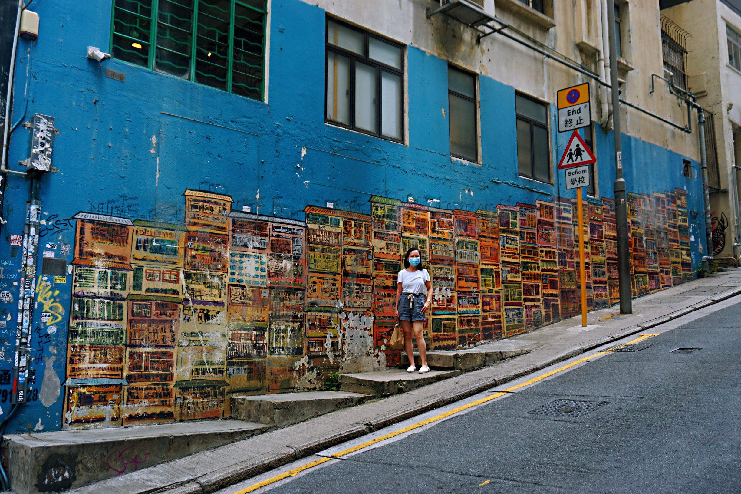  有人說，中上環就像濃縮了的香港，是旅人認識香港的一扇窗。大街小巷遍布古蹟、廟宇、畫廊、老字號、潮店、餐廳和酒吧……兼備古今中西，傳統與創意，繁華與寧靜。摩天大廈的巷弄裏，隱藏着一條色彩斑斕的壁畫線，「神出鬼沒」的壁畫塗鴉，不必刻意尋找，返工放工途中，就可能轉角遇到驚喜。而在港島的西南隅，西營盤的民居、黃竹坑的工廈間，也充滿了妙趣橫生的壁畫，成為大家朝夕相對的日常風景。圖為：中環嘉咸街階梯壁畫，以九龍城寨為背景
